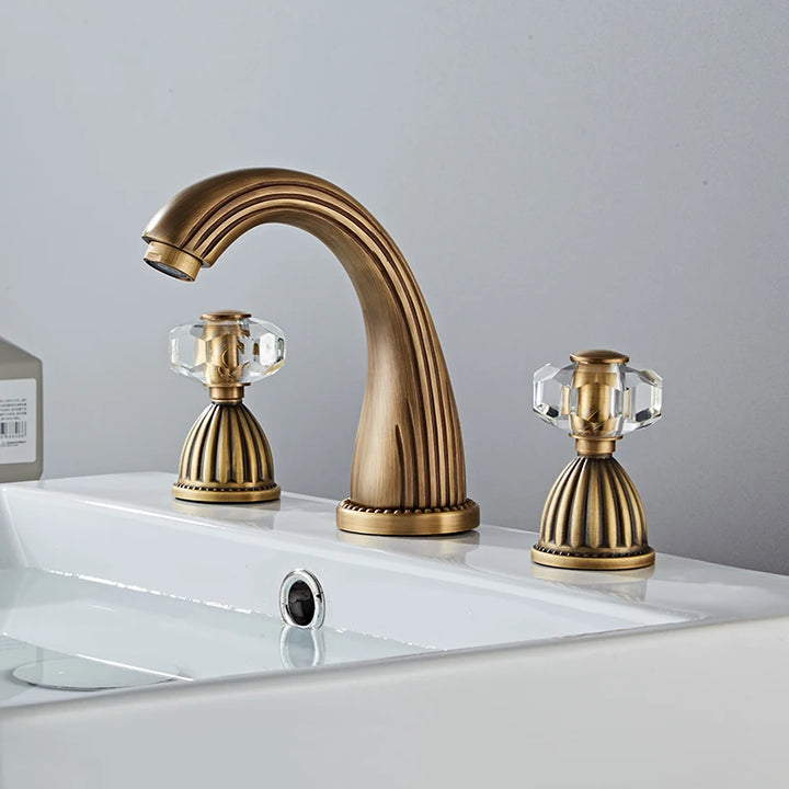 Dans une salle de bain épuré, il y a un lavabo blanc sur lequel est installé un robinet bronze de style rétro avec deux poignées déportées avec une pierre transparente sur chacune d'elles.