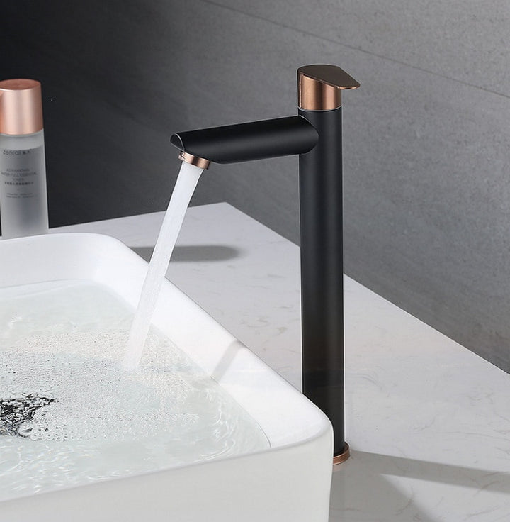 Un robinet lavabo bicolore avec eau qui coule dans un évier. Design minimaliste et contemporain avec courbes arrondies et finitions géométriques. Corps noir mat contrastant avec touches de doré brossé rose. Disponible en deux tailles pour salle de bain.