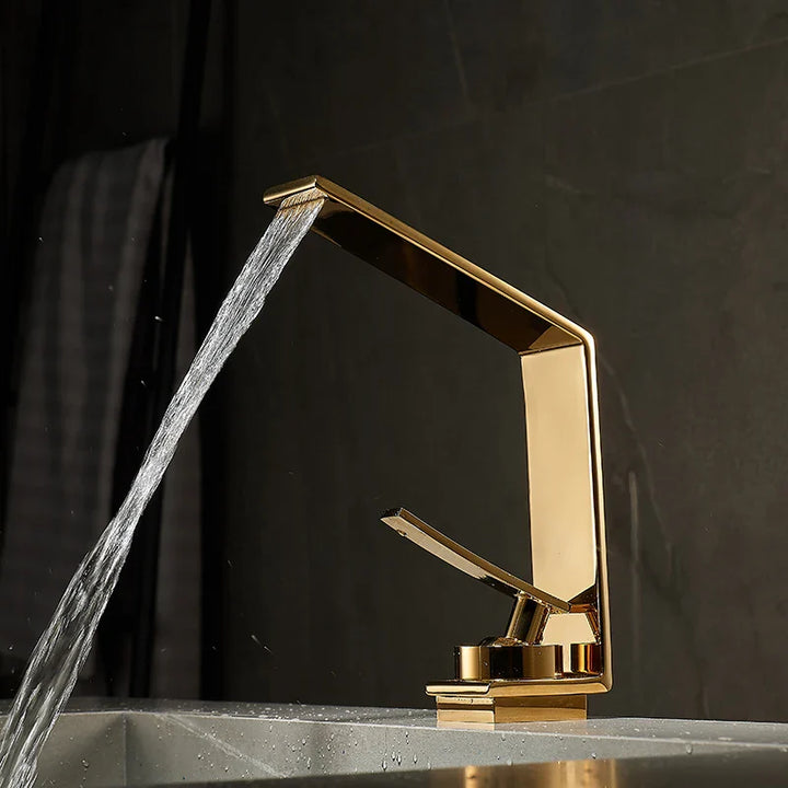 Dans une salle de bain au style moderne avec un mur en marbre noir, un robinet est posé sur un lavabo. Le robinet a une forme angulaire avec la poignée du mitigeur en dessous. Le robinet est doré brillant. 