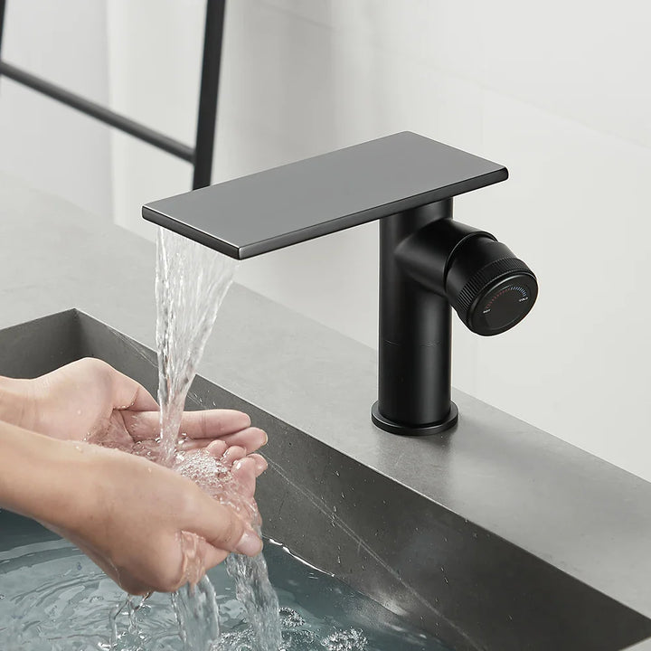 Dans une salle de bain, un lavabo est installé avec un robinet à bec plat et poignée sur le côté. Le robinet est noir mat. 