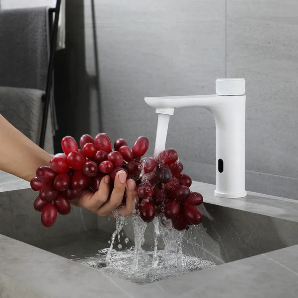 Dans une salle de bain en marbre, un robinet blanc est installé. Il est moderne avec une poignée ronde de mitigeur. Il est tactile. Une femme met une grappe de raisins sous l'eau.