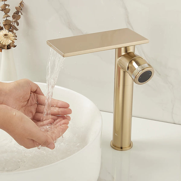 Dans une salle de bain, un lavabo est installé avec un robinet à bec plat et poignée sur le côté. Le robinet est doré brossé.